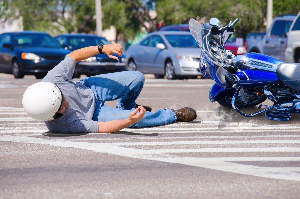 Motorcycle rider has wrecked and is laying in the road as his motorcycle goes sliding into the busy intersection. 