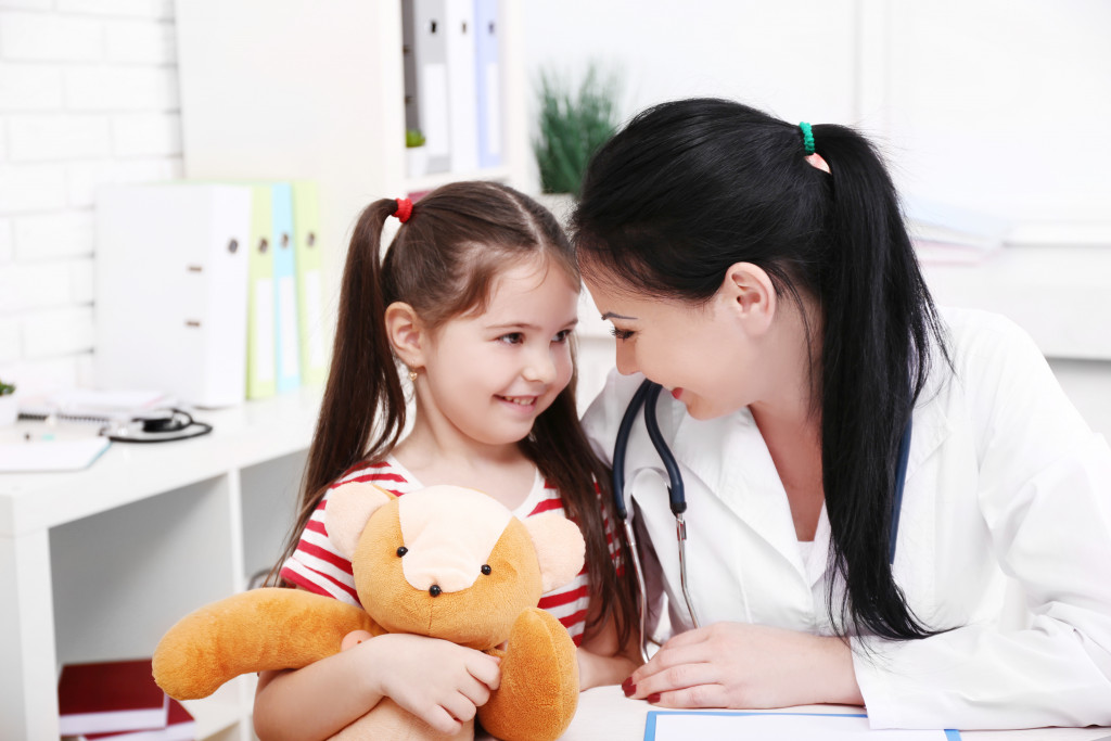 Young girl talking to a doctor while holding a teddy bear in a clinic.