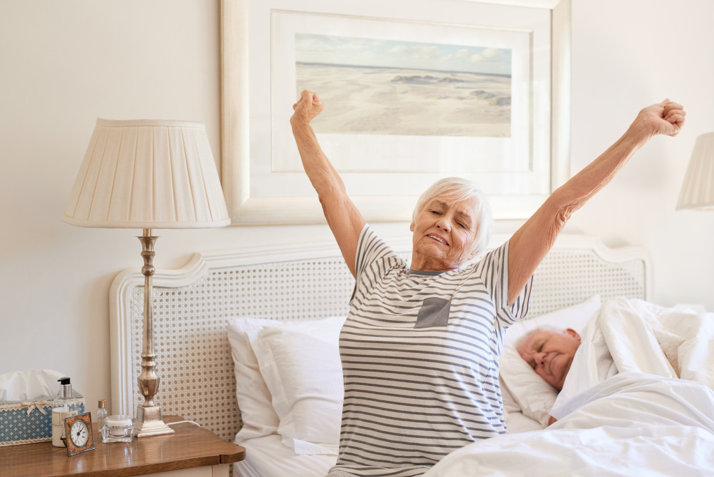 Senior woman sitting on her bed in the morning yawning with arms raised in a stretch with her husband sleeping next to her