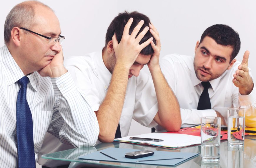 a man with his hands on his head sitting beside two co-workers in front of a table full of documents and glasses of water