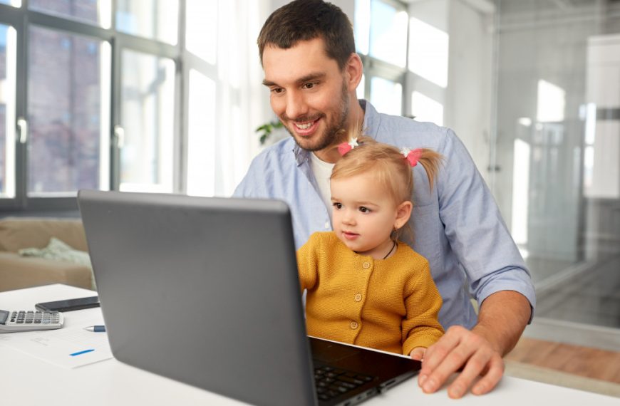 working father carrying daughter in lap while using laptop in office