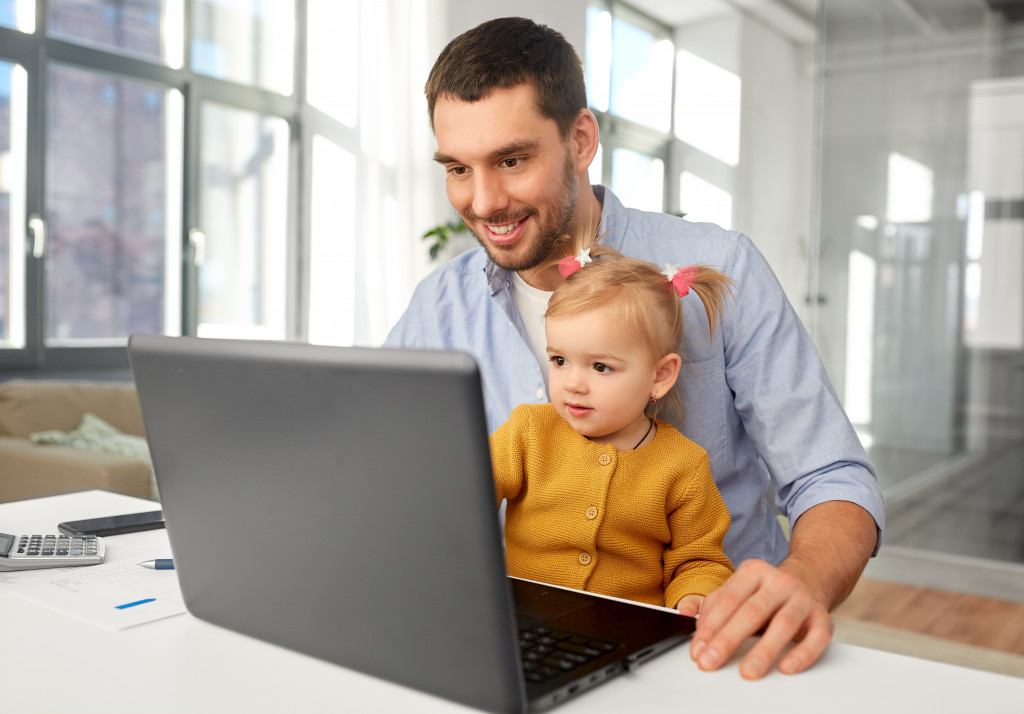 working father carrying daughter in lap while using laptop in office
