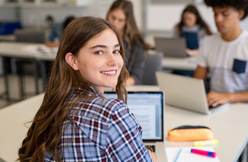 Portrait of smiling young woman student looking behind while using laptop