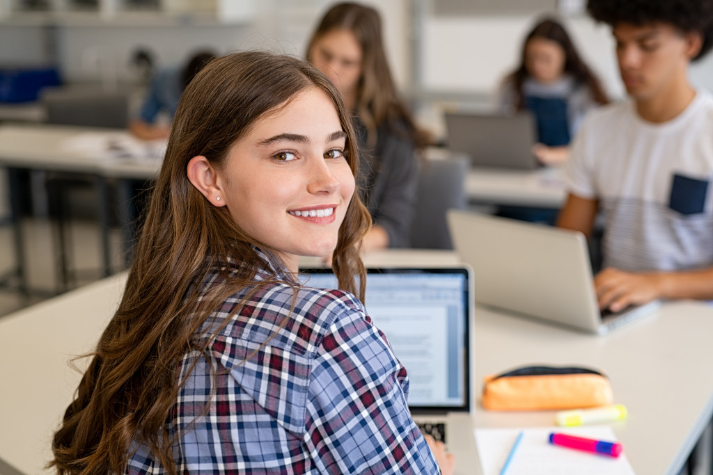 Portrait of smiling young woman student looking behind while using laptop
